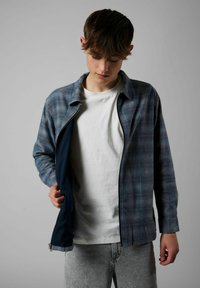 Sinine ja hall ruuduline lukkudega jakk pikkade varrukatega, millel on sile tekstuur ja tume siseserv, kantud valge T-särgi ja hallide teksade peal.