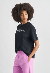 Μαύρο βαμβακερό t-shirt με λευκό λογότυπο «NIKE SHOX», σε συνδυασμό με ροζ φόρμα με μικρό ασημένιο λογότυπο Nike. Το μοντέλο κοιτάζει προς τα δεξιά.