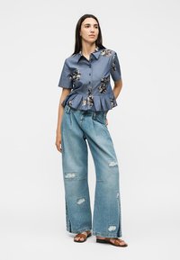 Femme debout portant une chemise peplum bleue à fleurs, un jean large déchiré et des sandales plates marron sur fond blanc.