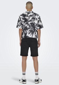 Camisa de manga corta con estampado de hojas de palma en blanco y negro, combinada con pantalones cortos negros enrollados. Usada con zapatillas deportivas en blanco y negro y calcetines a rayas.