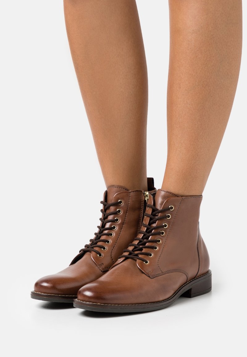 Paire de bottines marron en cuir à lacets portées sur des jambes nues, avec fermeture éclair latérale et petit talon empilé.