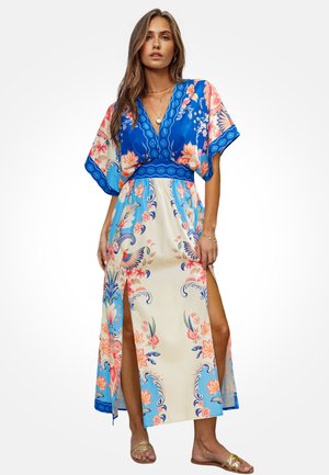 Vrouw die een maxi-jurk met korte mouwen draagt in blauw en crème met bloemenprint, met twee vooropensneden, staand met één hand langs haar zijde.