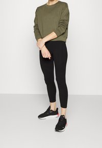 Olive green sweatshirt with a crew neck, black leggings with seams, and black sneakers with red accent. Relaxed fit, soft fabric.