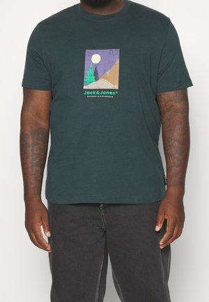 Mężczyzna w ciemnozielonym T-shircie z grafiką księżyca, drzew, wzgórz oraz napisem „Jack & Jones Designed in Copenhagen” na piersi.