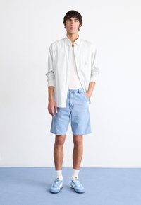 Camisa de botones blanca a rayas sobre una camiseta blanca sin mangas, combinada con pantalones cortos de mezclilla azul claro y zapatillas deportivas azules con acentos blancos.