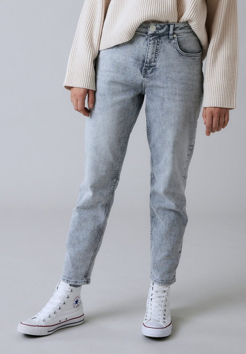 Personne portant un jean taille haute gris clair, des baskets montantes blanches et un pull côtelé crème à manches longues, debout sur un sol gris.