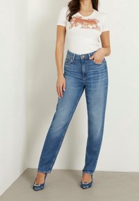 Biała koszulka z krótkim rękawem z kwiatowym wzorem, zestawiona z jasnoniebieskimi dżinsami o prostym kroju i niebieskimi butami na obcasie w wzory. Prosty, casualowy strój.