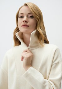 Cremfarbener Reißverschluss-Pullover aus Rippstoff, mit hohem Kragen und lockerer Passform, versehen mit sichtbaren Nahtdetails auf der Vorderseite.