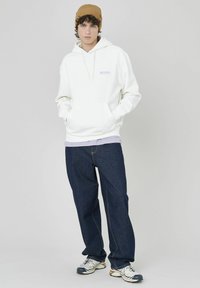 Sweatshirt à capuche blanche avec poche avant et texte violet, associé à un jean bleu foncé coupe ample et des baskets grises. Le modèle porte un chapeau marron.