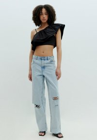 Schwarzes Oberteil mit einem Schulterträger und Rüschenstoff, kombiniert mit hellblauen verwaschenen Jeans mit Rissen und ausgefransten Kanten. Schwarze Sandalen sichtbar.