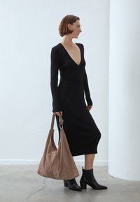 Czarna sukienka dzianinowa z głębokim dekoltem w kształcie litery V i długimi rękawami. Modelka trzyma miękką, brązową torebkę ze zamszu i ma na sobie czarne botki za kostkę na słupkowym obcasie.