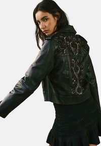 Giacca cropped in pelle nera con dettagli ricamati intricati sulla schiena, colletto a punte e chiusura con zip. Vestito nero arricciato visibile sotto.