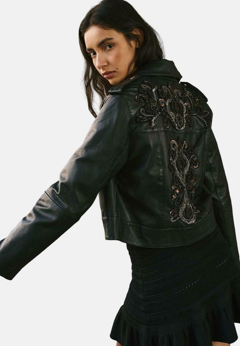 Giacca cropped in pelle nera con dettagli ricamati intricati sulla schiena, colletto a punte e chiusura con zip. Vestito nero arricciato visibile sotto.