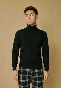 Pull à col roulé noir en maille, associé à un pantalon à carreaux noir et blanc. L'arrière-plan est d'un marron clair uni.