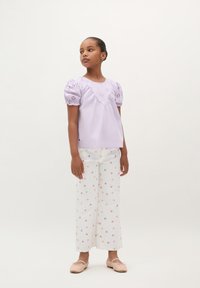 Lila kurzärmlige Bluse mit gesticktem Design und Puffärmeln, kombiniert mit weißen, weit geschnittenen Hosen mit floralen Mustern. Einfache rosa Schuhe.