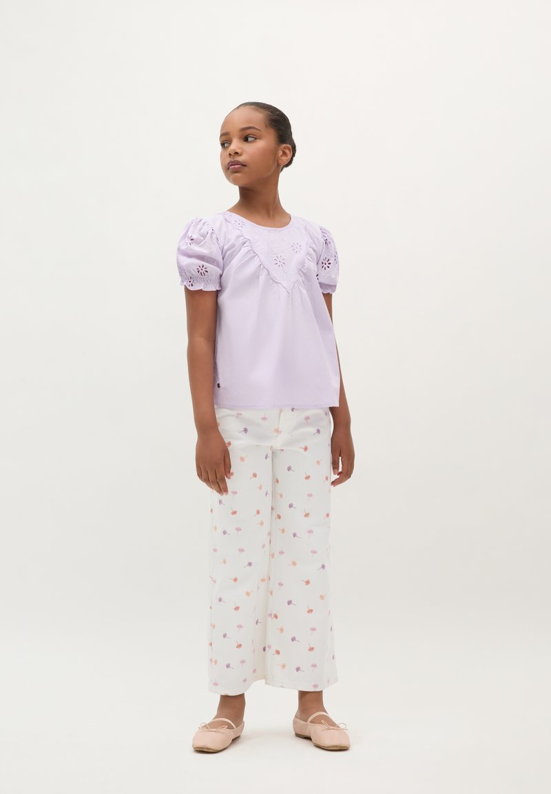 Lila kurzärmlige Bluse mit gesticktem Design und Puffärmeln, kombiniert mit weißen, weit geschnittenen Hosen mit floralen Mustern. Einfache rosa Schuhe.