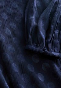 Dunkelblaue Bluse aus transparentem Stoff mit großen Polka-Dots und Puffärmeln. Verfügt über ein Knopfmanschetten-Detail zum Verschließen.