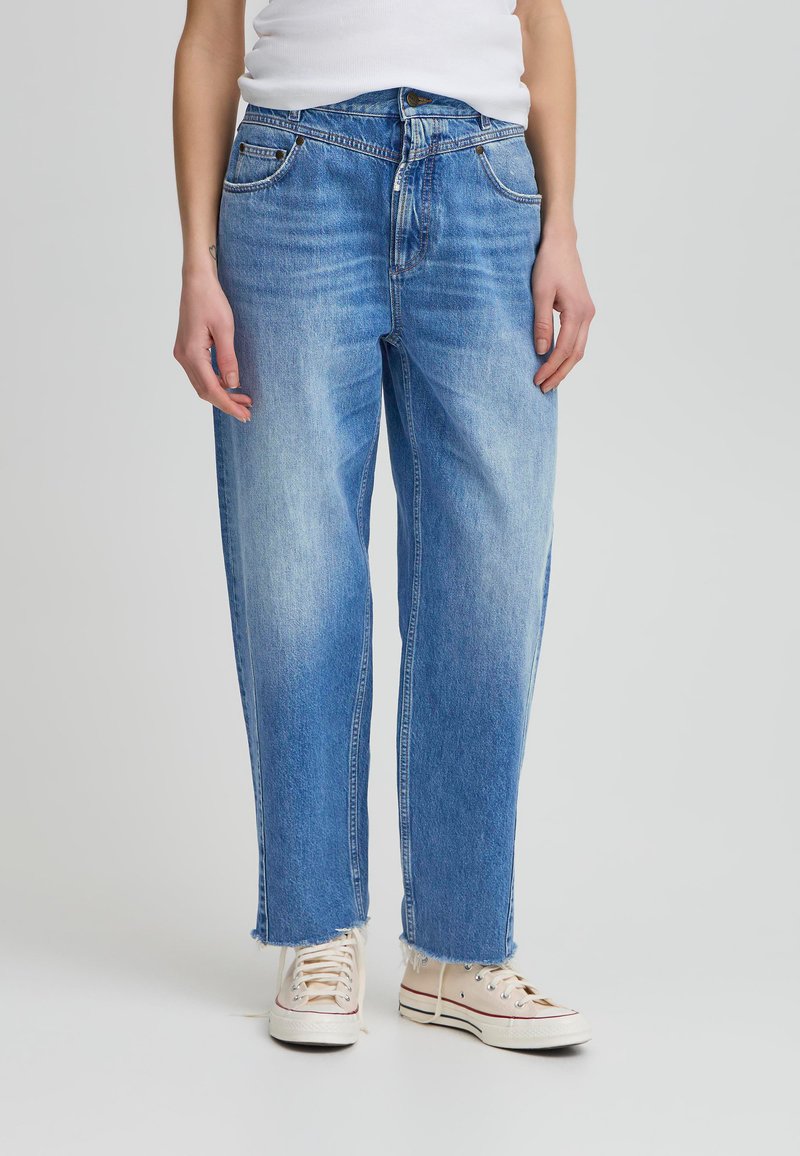 Gaiši zili platajā kājās džinsi ar izbalējušu izskatu, ar pogu aizdari, piecām kabatām un plīsušiem uzmaliņu galiem, kombinēti ar bēšām sporta apaviem.