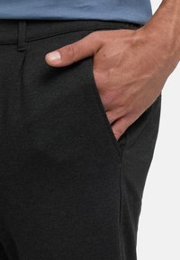 Die rechte Hand steckt in der Vordertasche einer schwarzen Hose, getragen mit einem blauen Hemd, das an der Taille sichtbar ist.