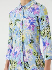 Svetlo modra cvetna srajca s tremi četrtinami rokavi, zapenjanjem spredaj in živahnimi vzorci rož v vijolični, zeleni in rumeni barvi.