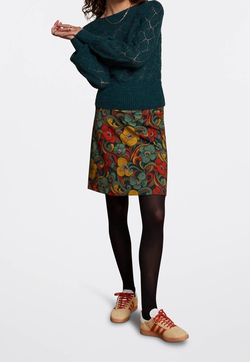 Turkoosiset neuleet, jossa on teksturoitu kuvio, kukkakuosiset hameet vihreässä, punaisessa ja keltaisessa, mustat sukkahousut ja beige tennarit punaisilla yksityiskohdilla.