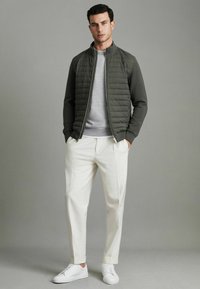 Veste bomber matelassée verte olive portée sur un pull gris clair, accompagnée d'un pantalon blanc plissé et de baskets blanches. Textures lisses et silhouette cintrée.