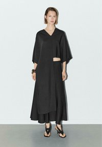 Kvinde iført sort asymmetrisk kjole med udsnit i taljen, sorte leggings, sorte strop sandaler og armbånd, stående op ad hvid væg.