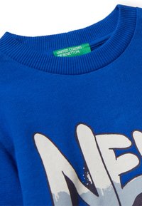 Suéter azul de cuello redondo con collar acanalado y texto gráfico blanco parcialmente visible, que presenta una etiqueta verde "United Colors of Benetton" en el interior.