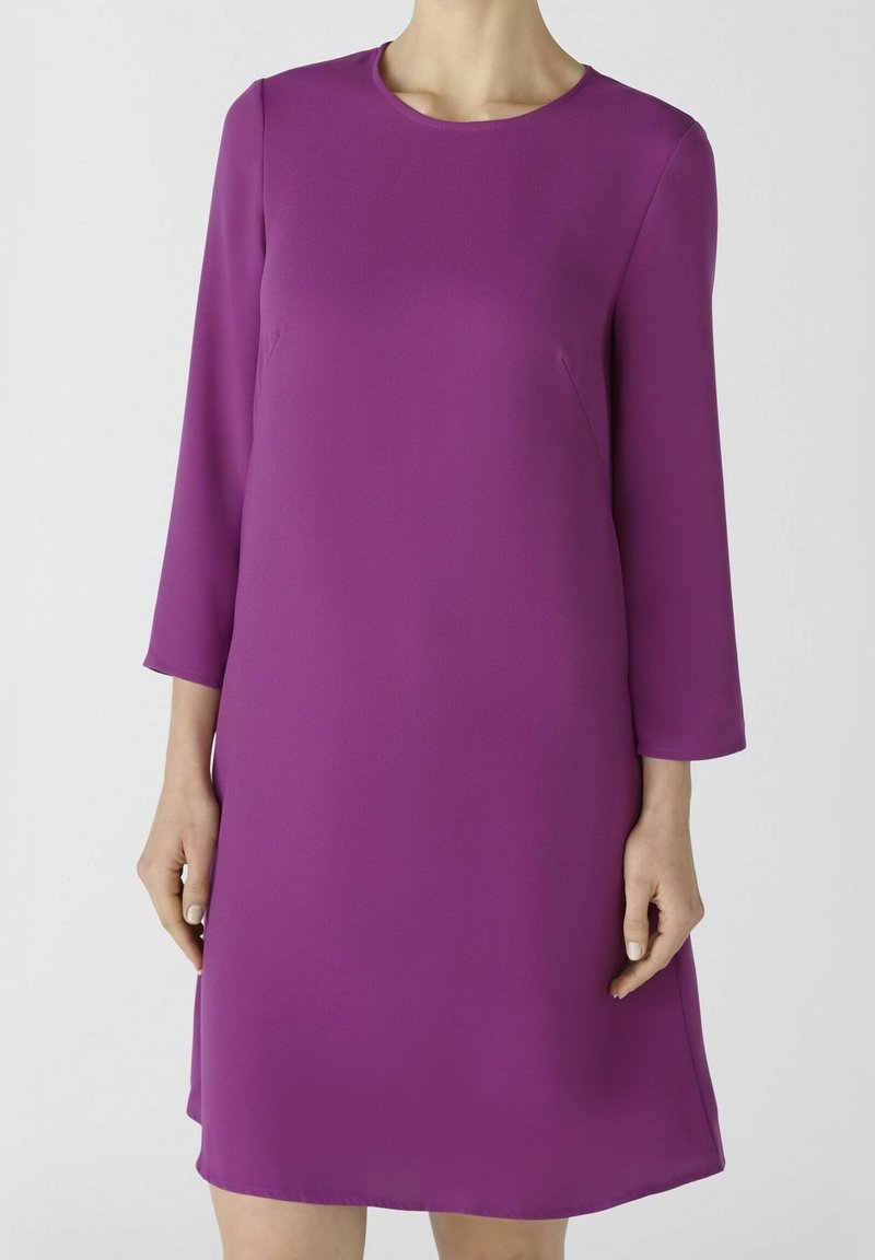 Femme portant une robe violette unie, longue jusqu’aux genoux, à manches longues, avec un col rond, sur un fond clair uni.