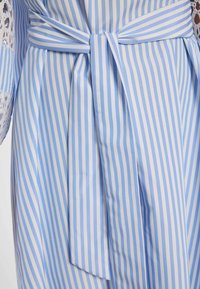 Blau-weiß gestreiftes Kleid mit vertikalen Streifen, einer gebundenen Schleife am Taillengürtel und seitlich sichtbaren Spitzensäumen an den Ärmeln.