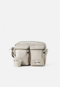 Bēšs Calvin Klein pārlocāms soma ar regulējamu siksnu, divas priekšējās kabatas ar rāvējslēdzēju un galvenā nodalījuma kabata ar rāvējslēdzēju uz baltas fona.