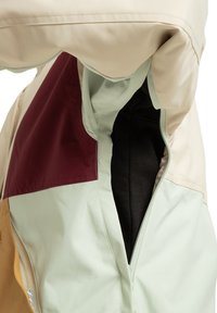 Bunda so zmesou krémových, svetlozelených, gaštanových a hnedých panelov; textúrovaný materiál; veľké bočné zipsy; a podšívkový interiér.