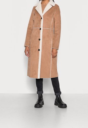 Femme portant un manteau en peau lainée beige longueur genoux avec bordure blanche, pantalon noir retroussé et bottes noires épaisses sur fond uni.