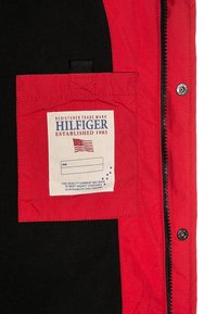 Rød jakke med sort kant, med et fremtrædende hvidt mærke med blå tekst, lynlåslukning og en lomme på venstre side.