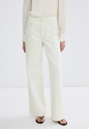 Persona vistiendo pantalones de tiro alto y pierna ancha de color blanco hueso con bordado floral sutil, combinados con una blusa crema transparente y sandalias negras.