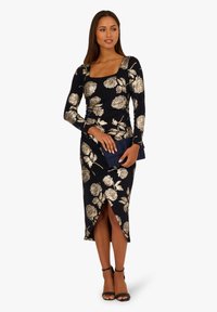 Abito nero con motivo floreale dorato, maniche lunghe, scollatura quadrata e spacco laterale. Indossato con una pochette blu navy e sandali neri con cinturini.