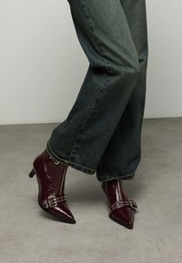 Bordeauxrode enkellaarsjes met een glanzende lakafwerking, een spitse neus, een kleine hak en een decoratieve gesp met metalen accenten. Jeans met een losse pasvorm.