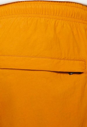 Oranssit shortsit, joissa on joustava vyötärö, takaosassa litteä tasku ja teksturoitu kangas. Vähäistä ompelutaitoa on näkyvissä.
