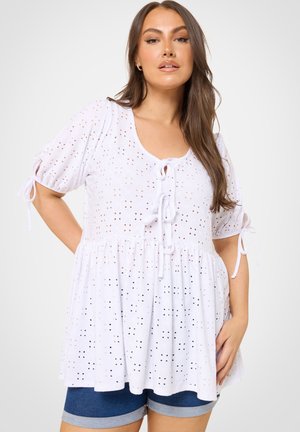 Kobieta ubrana w białą bluzkę z krótkimi rękawami z ażurowym wzorem, wiązaną z przodu i na rękawach, w połączeniu z podwiniętymi niebieskimi szortami dżinsowymi.