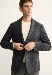 Blazer grigio a spina di pesce con rever, chiusura a un bottone e tasche frontali, sovrapposto a un maglione a collo rotondo color crema.