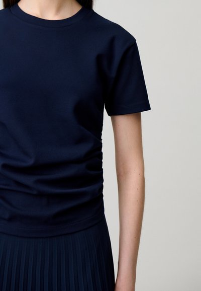 Persona che indossa una maglietta blu scuro a maniche corte semplice e una gonna plissettata scura abbinata su uno sfondo neutro.