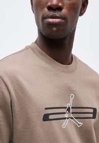 Κοντινό πλάνο ενός άνδρα που φοράει μπεζ φούτερ με ασπρόμαυρο γραφικό ενός παίκτη μπάσκετ που πηδά με τη μπάλα.