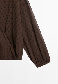 Braune Langarmbluse mit schwarzen Polka-Dots, mit voluminösen Ärmeln und gerafften Bündchen, flach auf weißem Hintergrund ausgelegt.