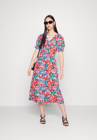 Vestido midi floral com padrões em vermelho, azul e verde, com mangas bufantes e cintura ajustada, combinado com sandálias pretas de tiras e óculos de sol.