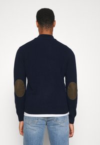 Granatowy sweter z prążkowanym kołnierzem i brązowymi zamszowymi łatami na łokciach, wykonany z miękkiego dzianego materiału, połączony z jasnoniebieskimi dżinsami.