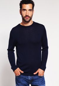 Mörkblå stickad tröja med rund halsringning, långa ärmar och ribbade muddar. Tyget verkar mjukt och lätt, kombinerat med blå jeans.
