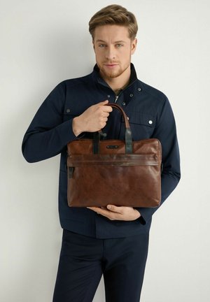 Młody mężczyzna w granatowej kurtce trzymający brązową skórzaną teczkę z czarnymi uchwytami, stojący na tle jasnego, jednolitego tła.