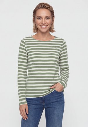 Lächelnde Frau mit kurzen blonden Haaren, die ein grün-weiß gestreiftes Langarmshirt und blaue Jeans trägt, steht mit einer Hand in der Tasche.