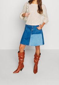 Woman wearing beige knit sweater, blue patchwork denim skirt, brown slouchy leather boots, and holding snakeskin-print shoulder bag.