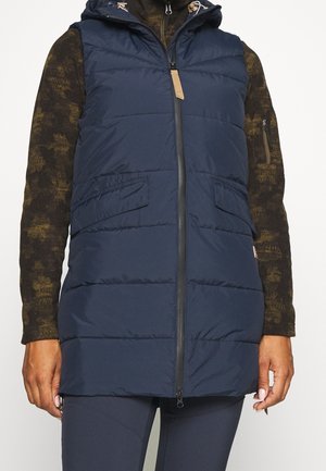 Personne portant un gilet sans manches matelassé bleu marine avec une fermeture éclair sur le devant, par-dessus un haut à manches longues sombre à motifs, visible du cou aux hanches.